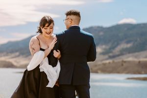 Elegant couple in formal attire pose against mountain lake backdrop.