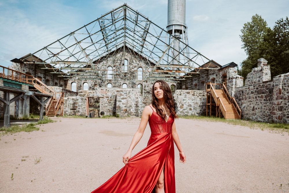 Elegant figure in flowing red dress poses gracefully among rustic stone ruins and greenhouse structures.