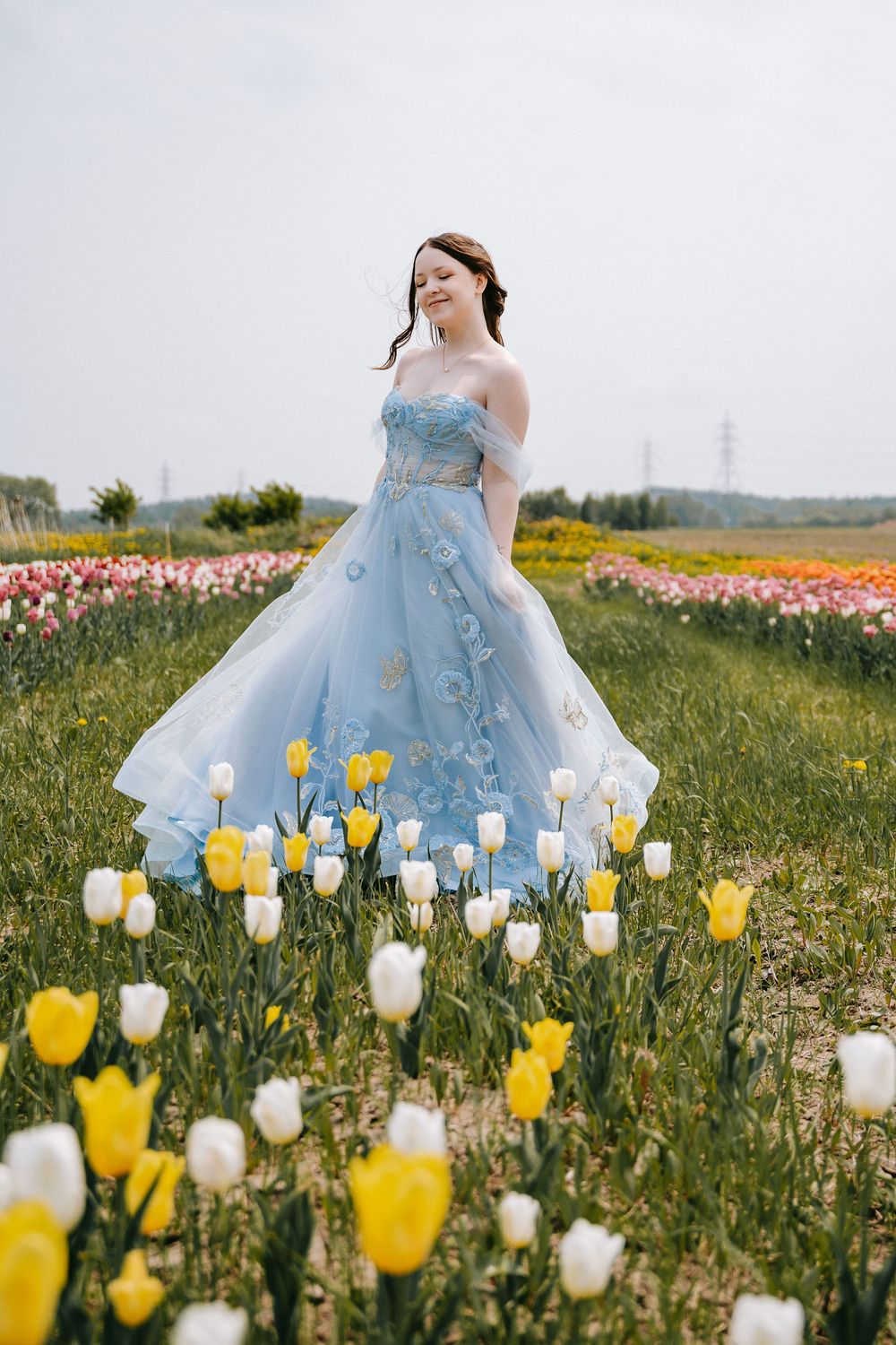 A light blue ballgown dress stands out among vibrant yellow and white tulips in a springtime field.