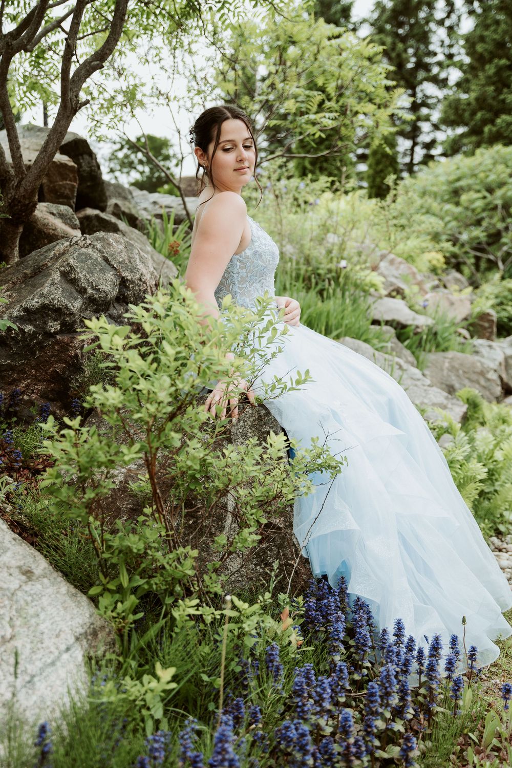 A light blue ballgown flows elegantly amongst purple wildflowers and green foliage in a natural outdoor garden setting.