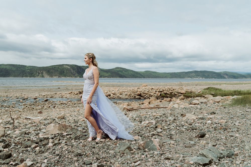 A person in a flowy white dress walks along a rocky beach shoreline on a cloudy day.