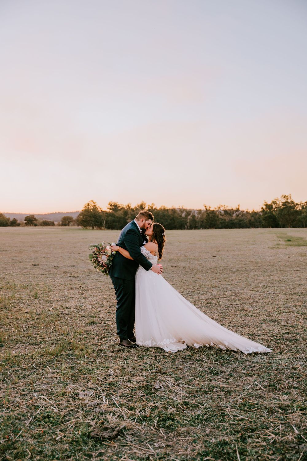 Newlyweds share a romantic moment in a rustic field during sunset.