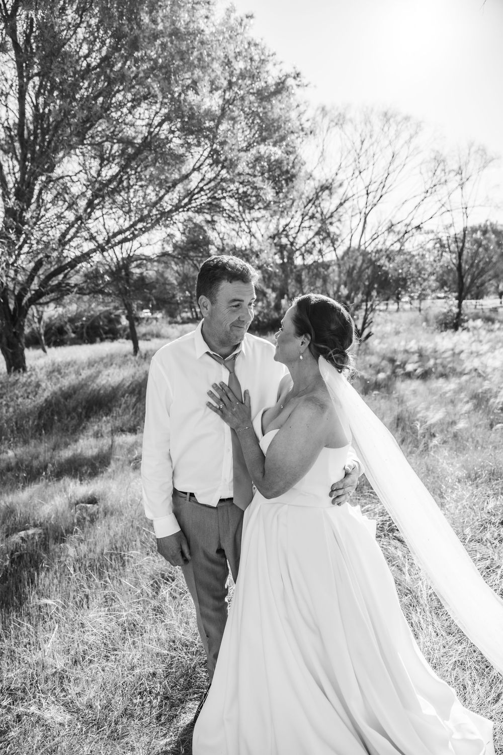 A couple in wedding attire pose together in a grassy field with trees in the background.