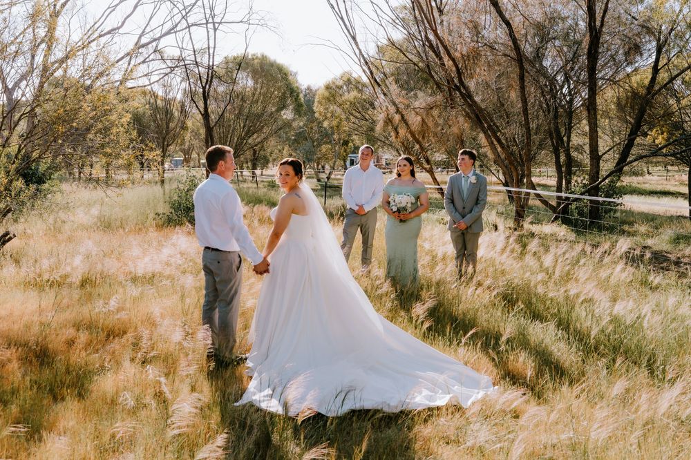 Wedding couple walking through tall grass with guests visible in the background.