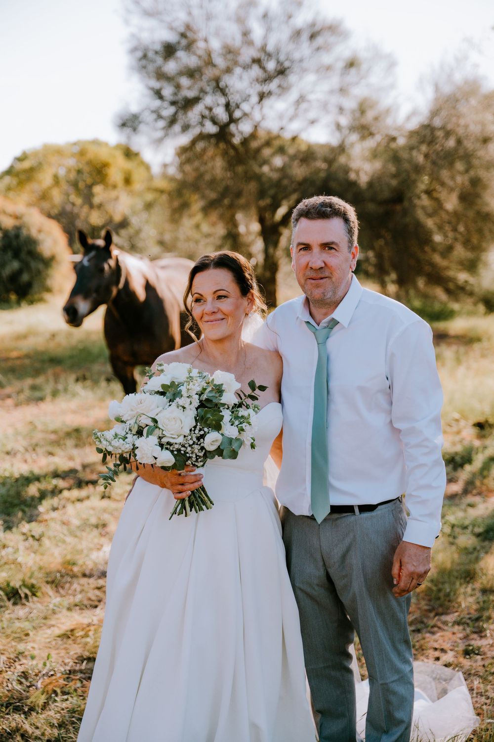 Wedding couple posing with white floral bouquet in an outdoor rustic setting.