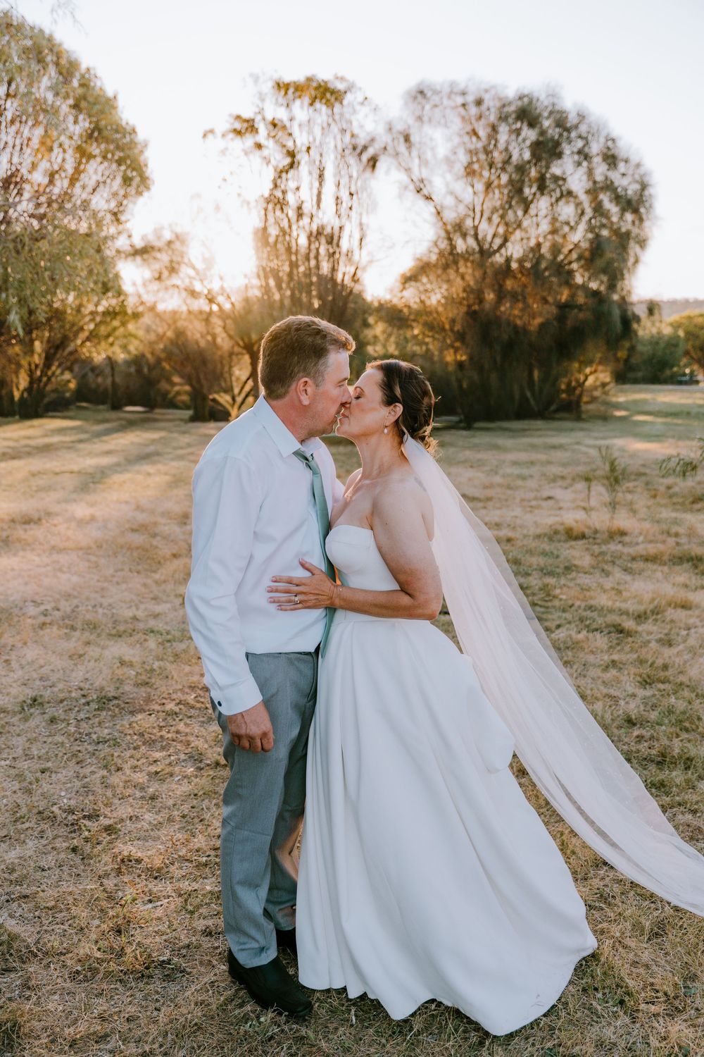 A romantic sunset wedding photo of a couple embracing in a field with golden light and flowing veil.