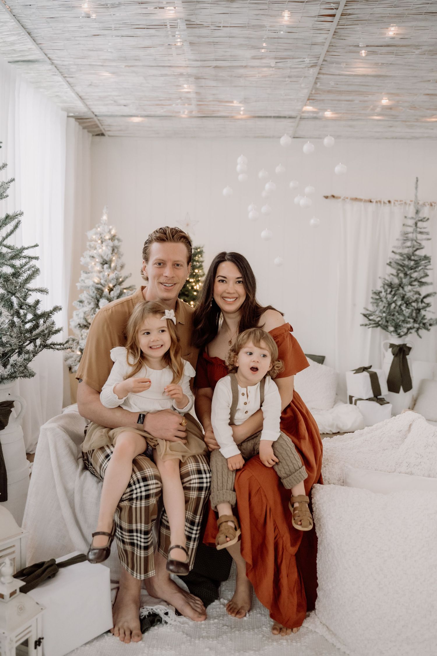 A family poses together in a cozy white holiday setting with decorated Christmas trees and twinkling lights.