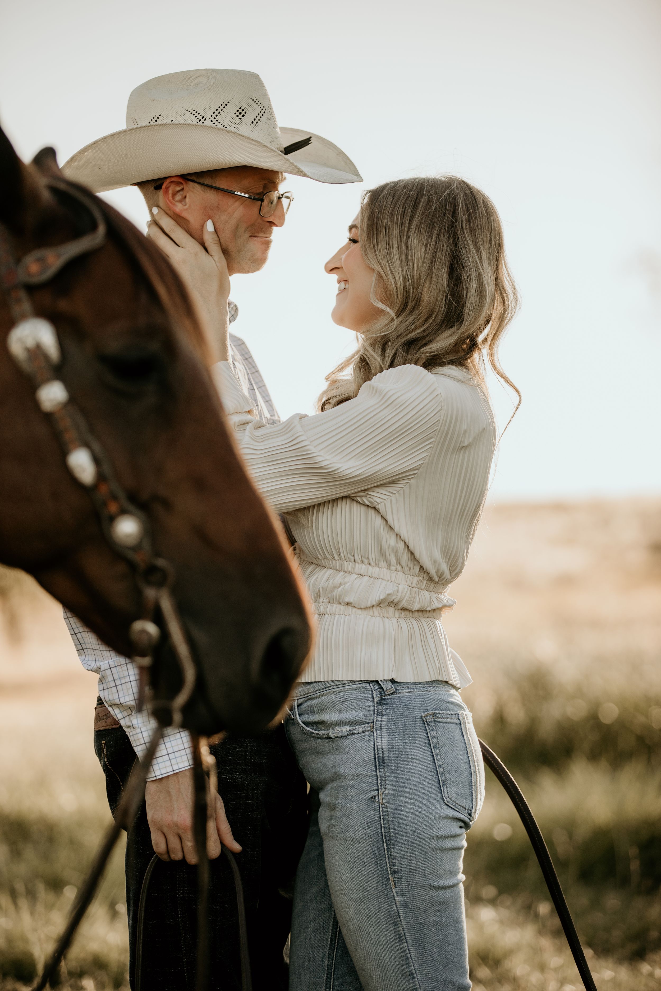 A romantic couple embraces next to a brown horse during a golden hour photoshoot in the countryside.