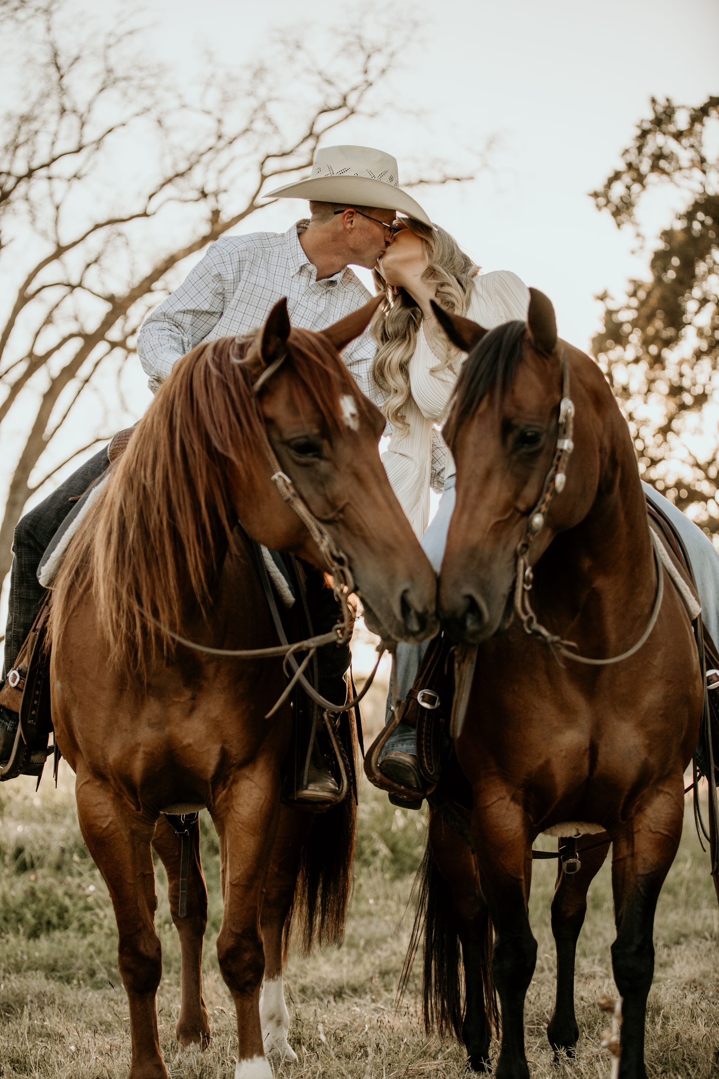 A romantic couple on horseback pose with their two brown horses in a rustic outdoor setting at sunset.