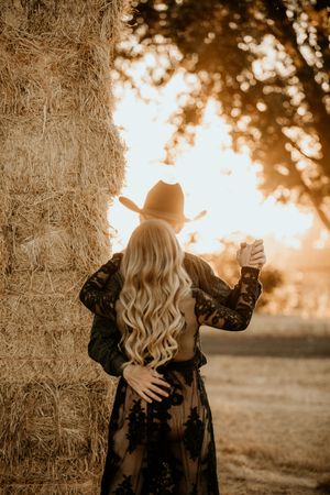 Two figures embrace against a rustic hay bale wall in golden sunset light, wearing elegant black lace clothing.