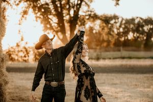 Romantic couple embraces in a golden field during sunset while wearing dark casual clothing.