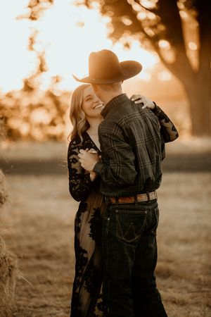 A romantic sunset portrait of a couple embracing outdoors, with a cowboy hat and warm golden lighting through the trees.