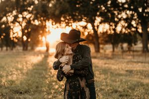A couple embraces in a golden sunset field while wearing western style clothing and a cowboy hat.