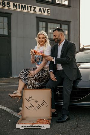 Couple sharing pizza while sitting on cardboard boxes on an urban sidewalk.