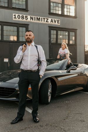Stylishly dressed man in formal attire poses near parked cars on urban street with '1085 Nimitz Ave' address visible.