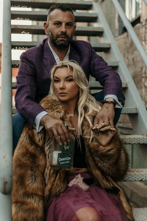Two people in formal attire posing on industrial metal steps with brown fur coats.
