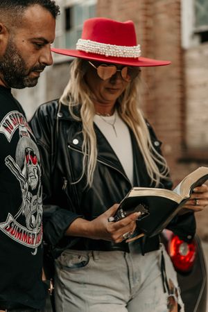 A couple standing close together look at their phones while wearing black leather and a red hat outdoors.