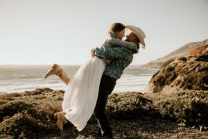 A romantic couple embracing on a coastal cliff with ocean and rocky shoreline in the background during golden hour.