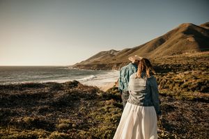 Couple embraces on coastal cliff overlooking rugged California shoreline at sunset with mountains in background.