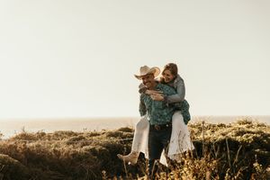 Couple embracing in warm golden sunlight on grassy hilltop during sunset with mountains in background.