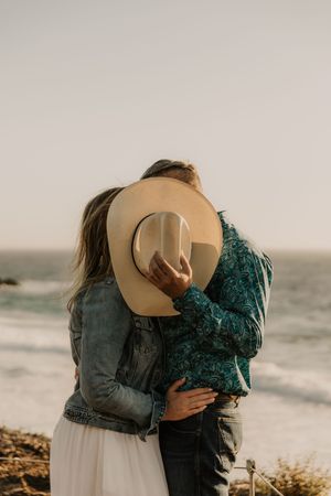 A romantic embrace between a couple at the beach during sunset, with one wearing a tan hat and a green sweater.