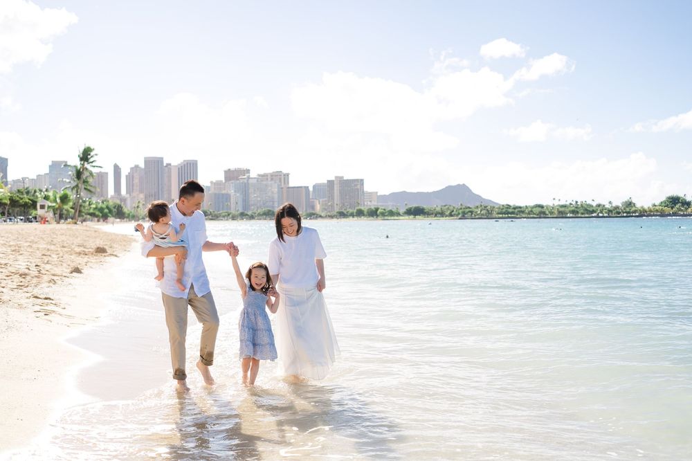 Family in white and blue outfits walks together on a scenic Hawaiian beach with ocean and mountains in background.