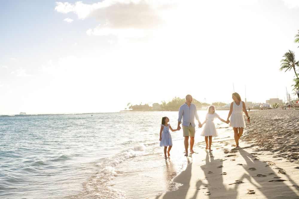 Family silhouettes walking together along a tropical beach shoreline at sunset with palm trees in background.