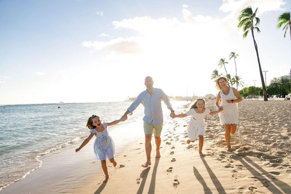 A family holds hands and walks together along a sandy beach at sunset with palm trees in the background.