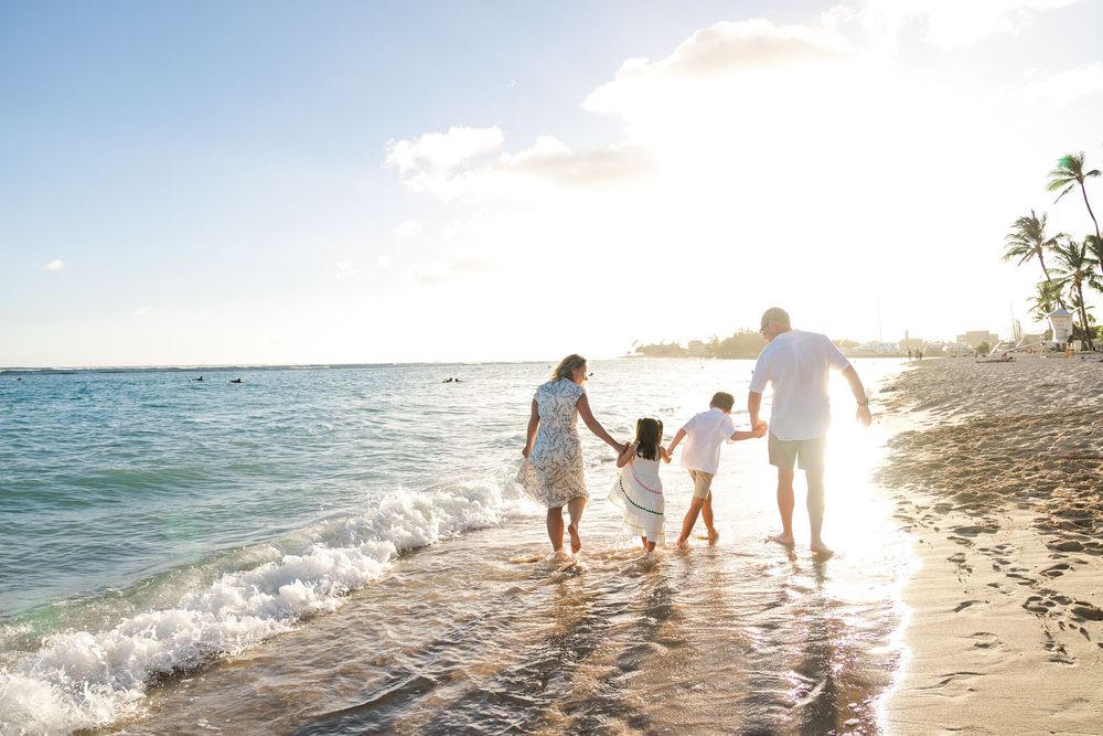 A family walks together along a tropical beach shoreline at sunset with waves lapping at their feet.