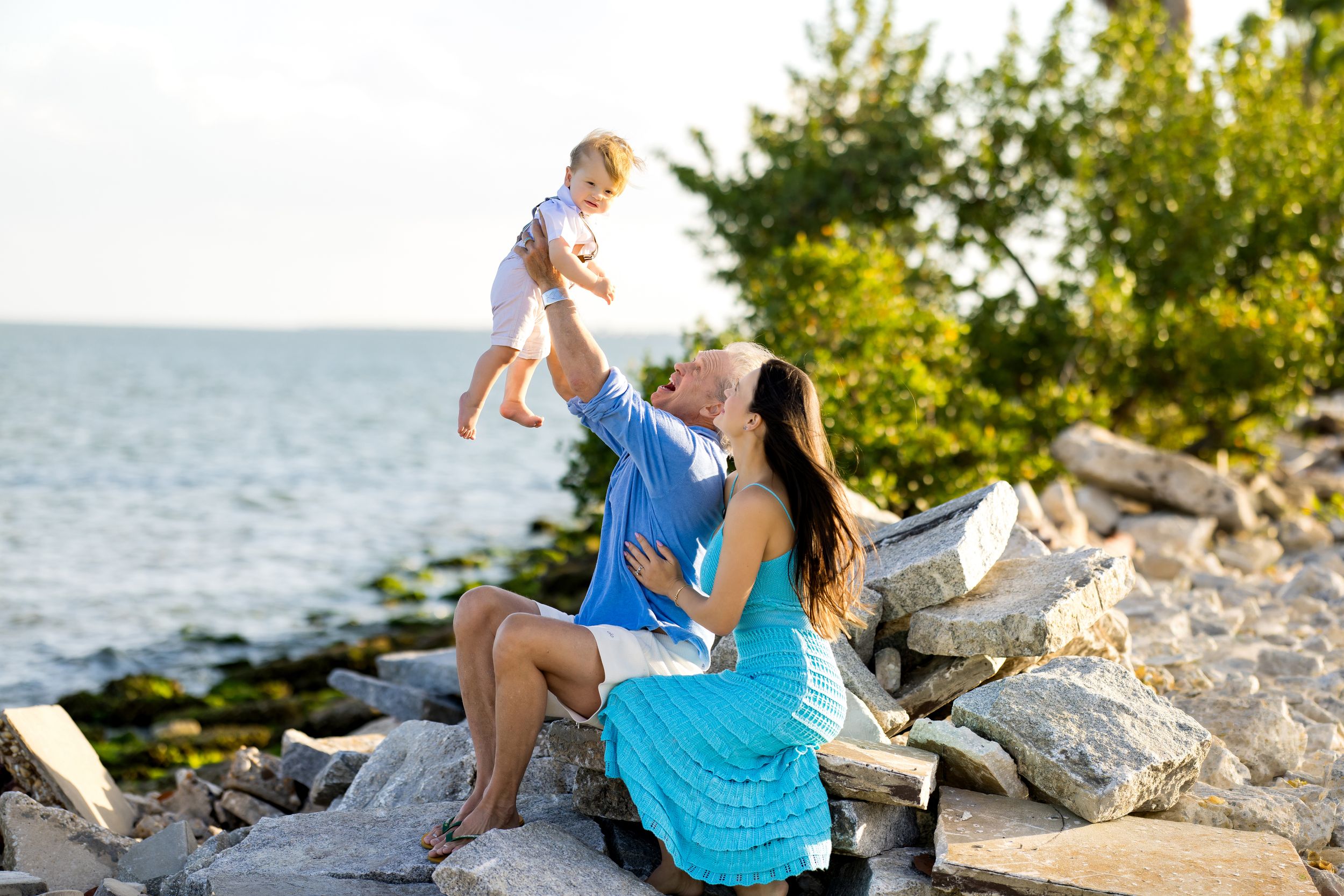 Family enjoying a sunny day on rocky shoreline, father lifting baby while mother sits in turquoise dress near calm water.