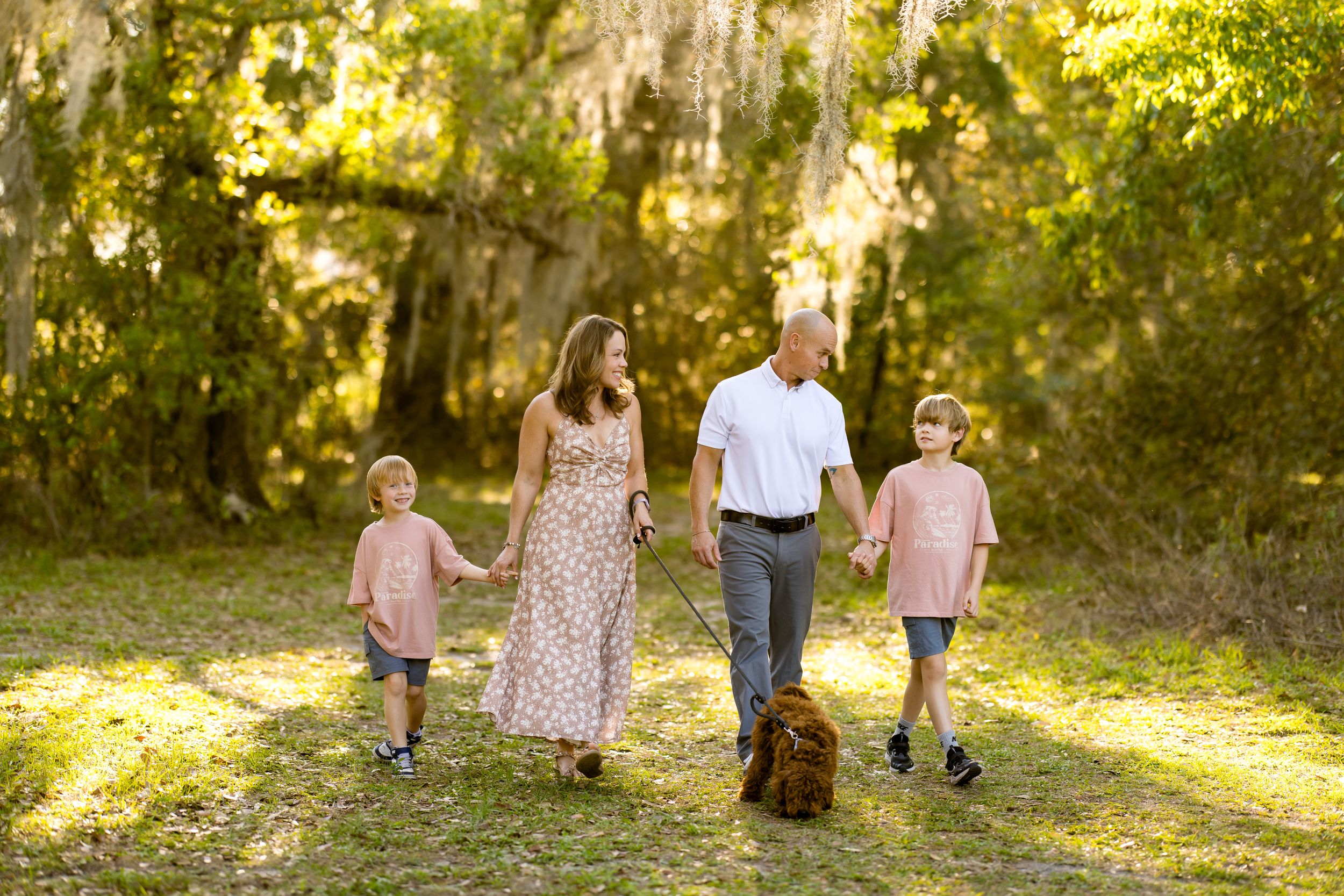 Family of four walking with dog on sunny forest path, parents holding hands with two boys in matching pink shirts.