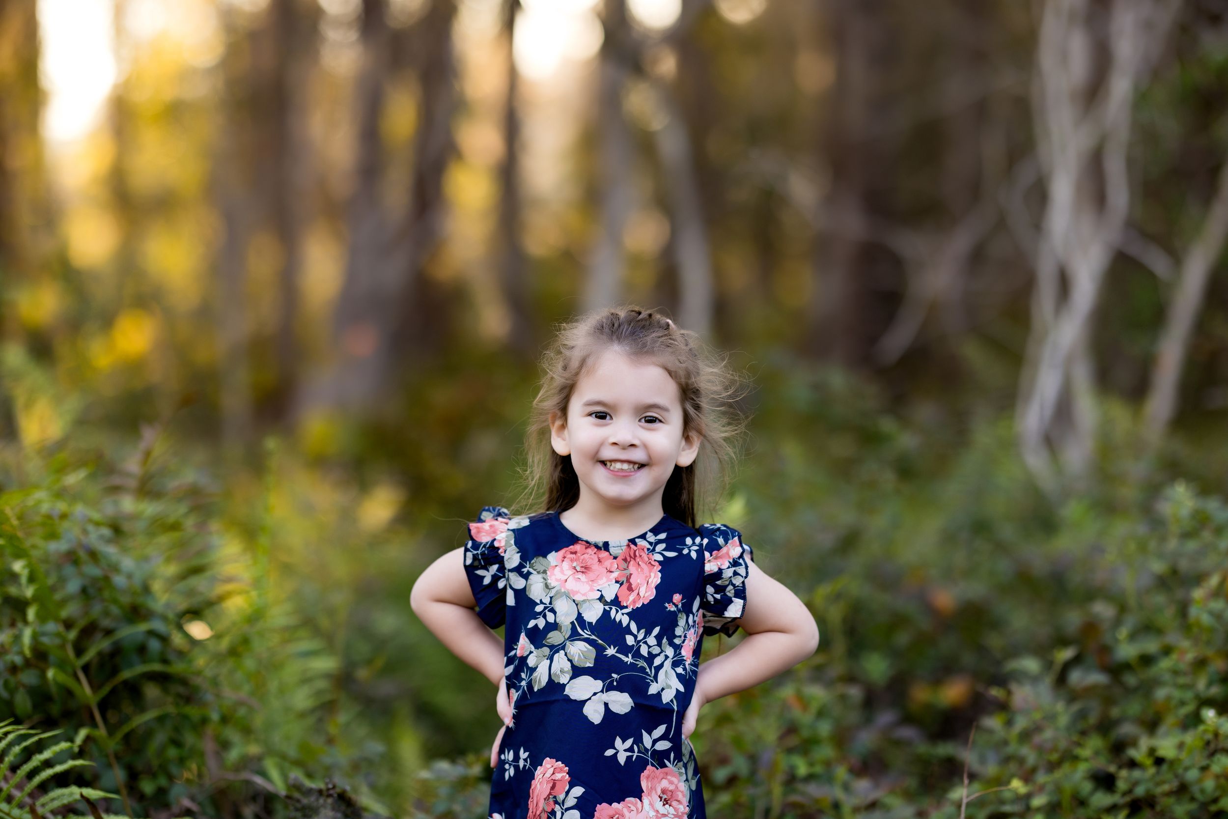 Child in floral dress poses against blurred forest background at sunset.