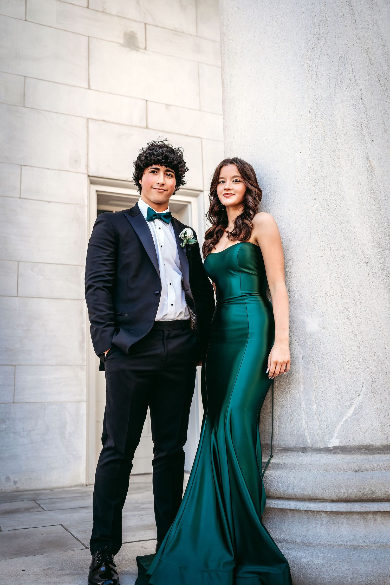 Formal couple in tuxedo and emerald green gown posing against white stone wall.