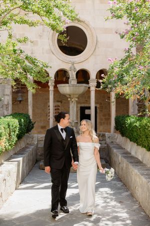 Couple in formal wedding attire walking through a beautiful stone archway corridor with flowering vines.