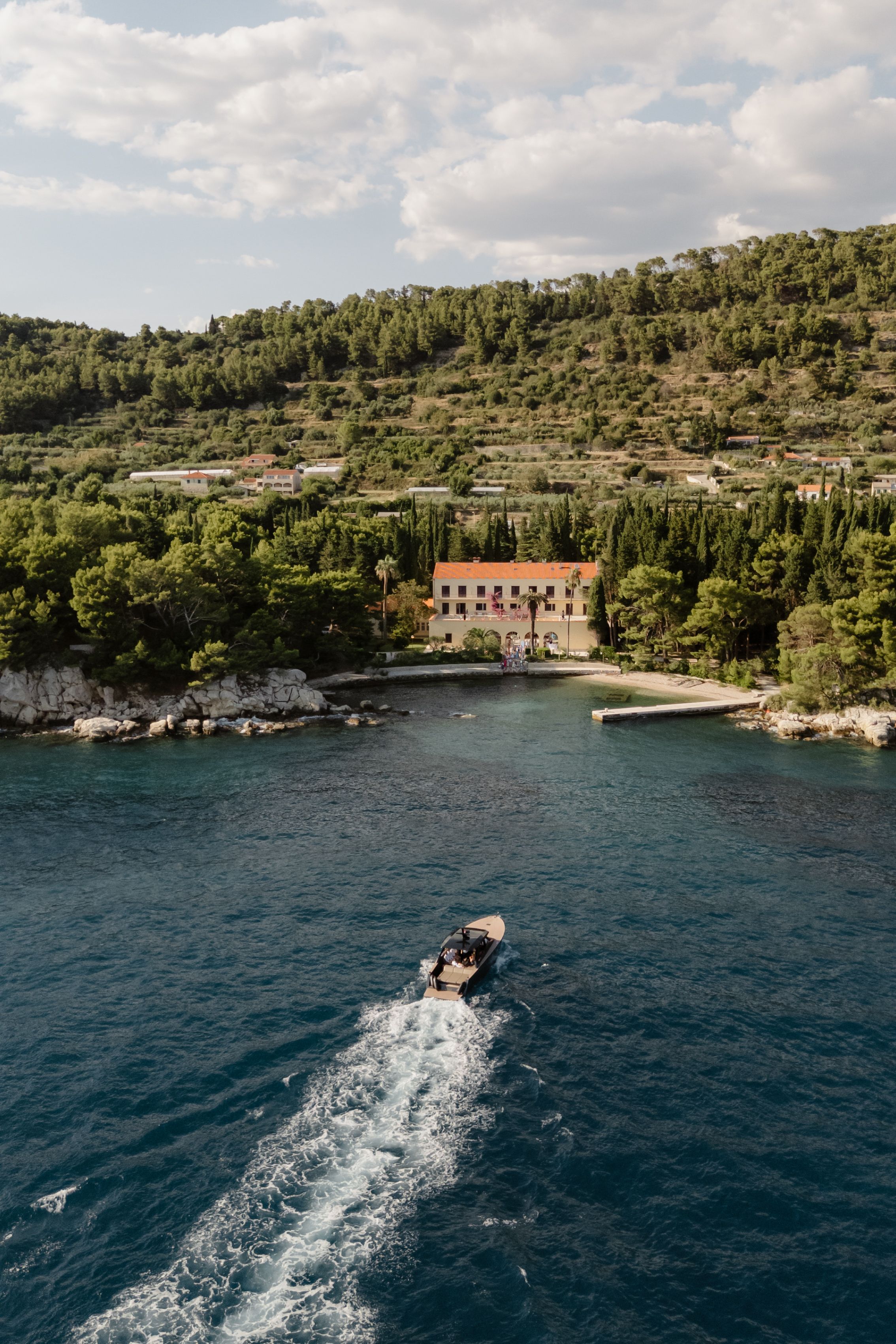 Aerial view of a boat creating wake in blue Mediterranean waters near a coastal villa and rocky shoreline.