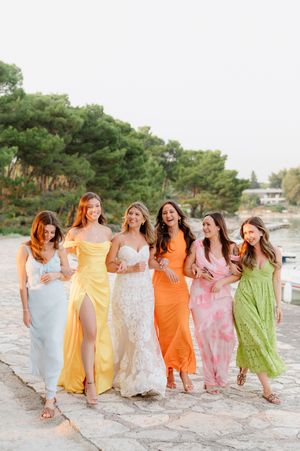 A group in colorful flowy dresses laughs together while walking along a sandy beach at sunset.