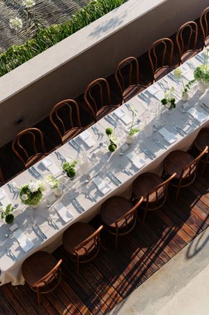 Aerial view of elegant long dining table with white linens, green floral centerpieces, and brown bentwood chairs outdoors.