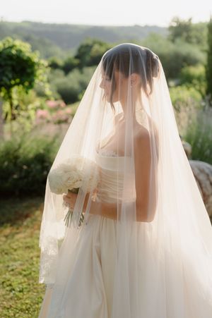 Bride in white gown and flowing veil holds white bouquet in lush garden setting.