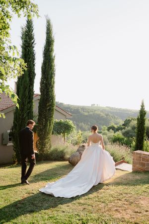 Bride in flowing white gown with train walks with groom in black suit through lush Tuscan garden with cypress trees.