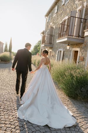 Bride and groom walking hand in hand on cobblestone path beside lavender bushes at a rustic stone villa.