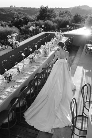 Bride in elegant ballgown wedding dress stands beside long reception table on outdoor terrace, black and white photo.