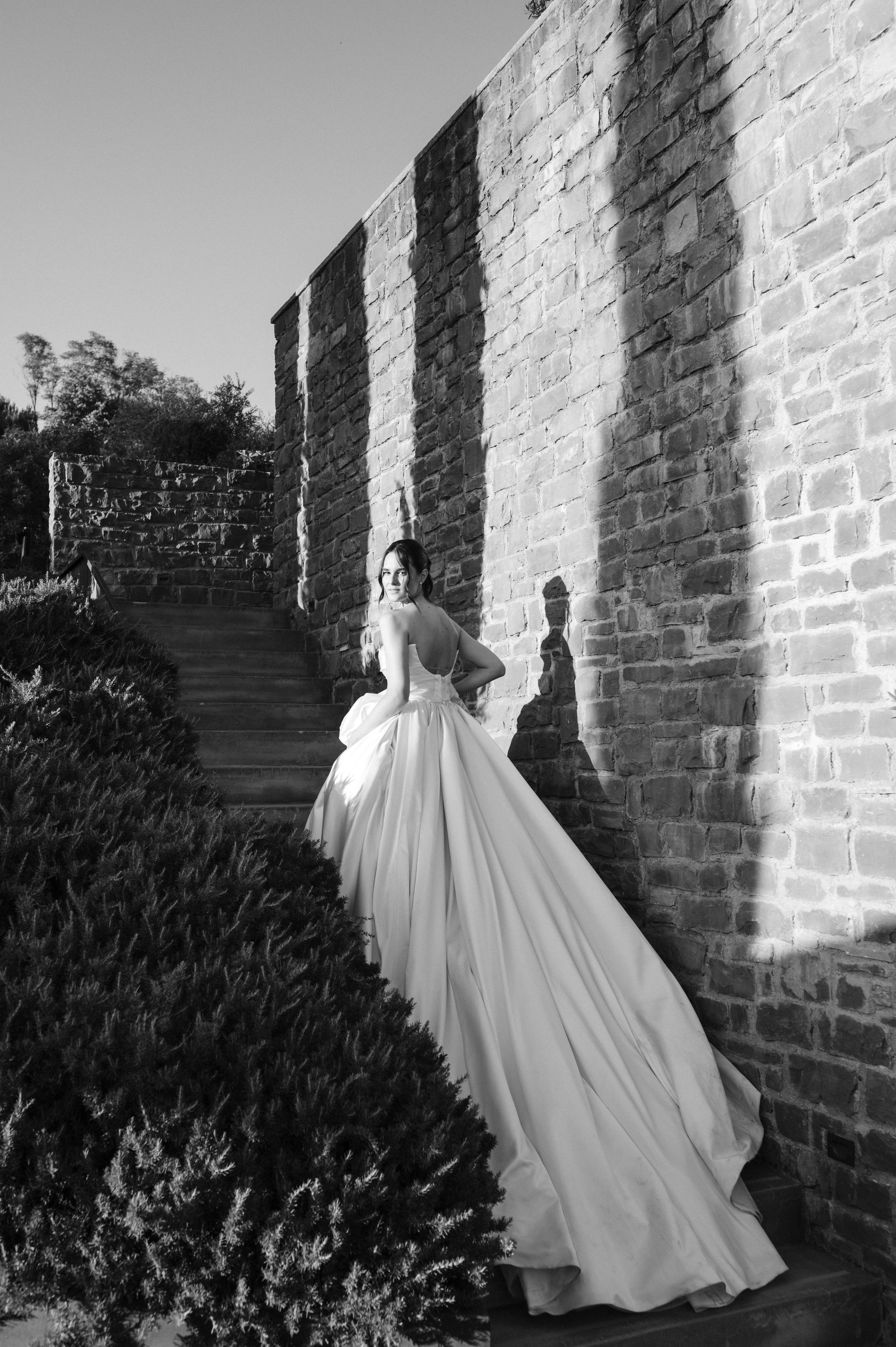 Black and white photo of a bride in a flowing ball gown standing against a rustic stone wall with dramatic shadows.