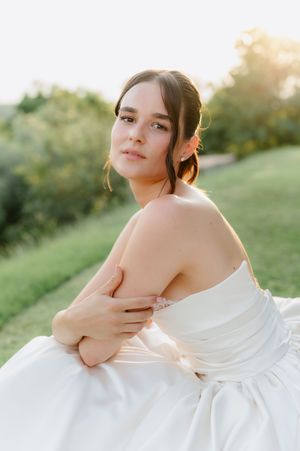 Young bride in strapless white gown sits outdoors in golden sunlight, arms crossed, gazing softly at camera.