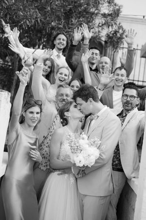 Wedding party celebrates with raised hands as the couple shares a romantic kiss while holding a white bouquet.