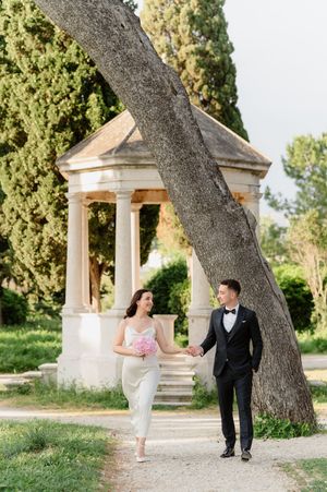 A couple in formal wedding attire playfully interacts beneath a curved tree trunk near classical stone gazebos.