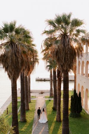 A couple walks along a path lined with tall palm trees beside a white Mediterranean-style building.