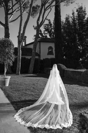 A flowing white wedding veil trails elegantly across a garden lawn with a Mediterranean-style building in the background.