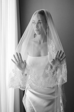 Black and white photo of a bride in a white gown and lace veil, hands raised, posing elegantly by a window.