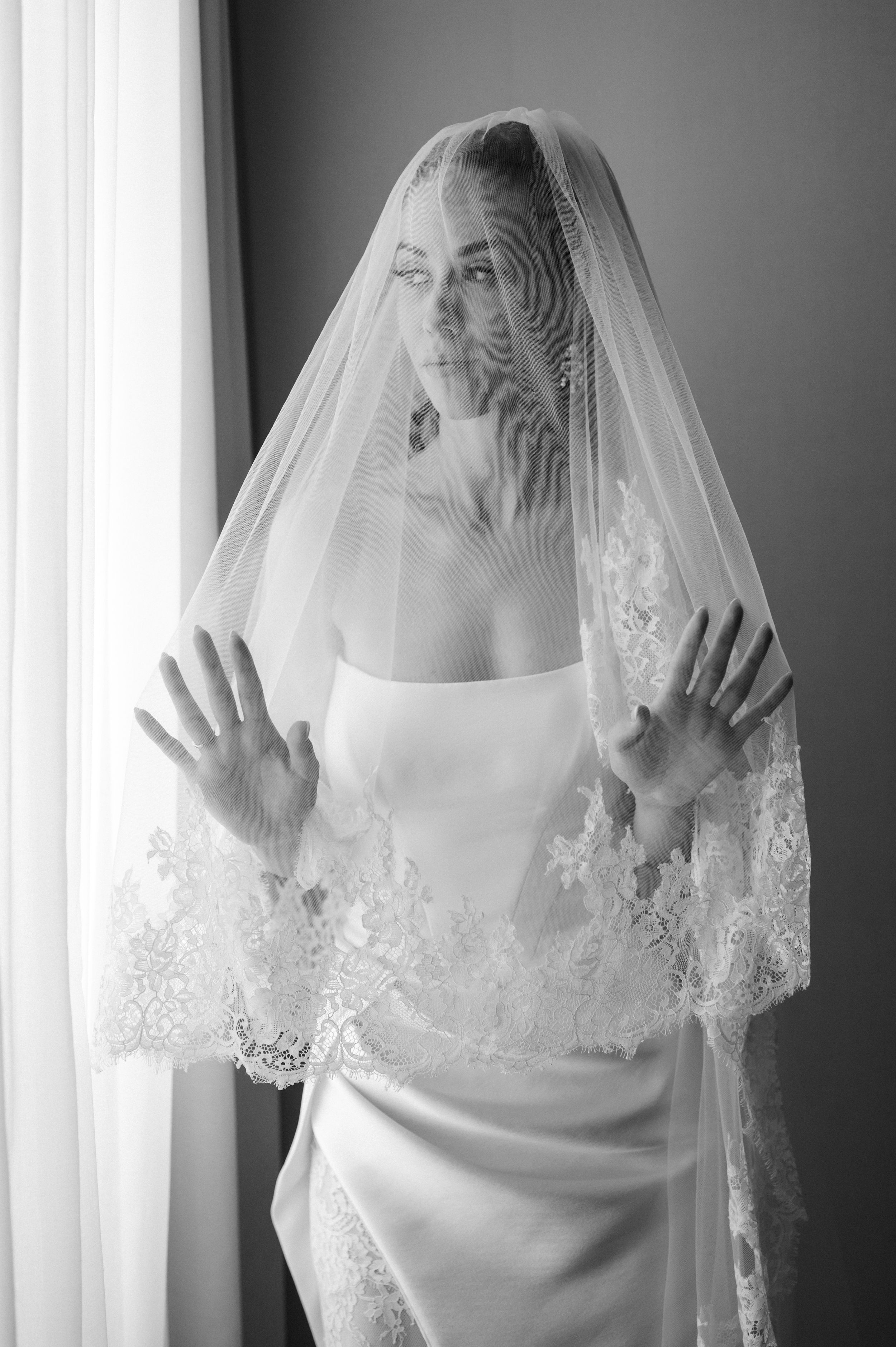 Black and white photo of a bride in a white gown and lace veil, hands raised, posing elegantly by a window.