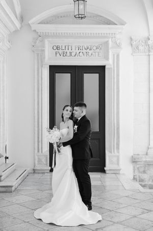 Black and white wedding photo of bride and groom embracing before ornate classical doorway with Latin inscription.